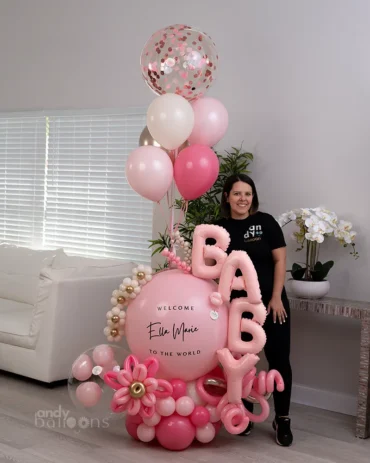 Glamorous pink and gold baby girl balloon bouquet with personalized "Welcome to the world" message and confetti-filled topper.