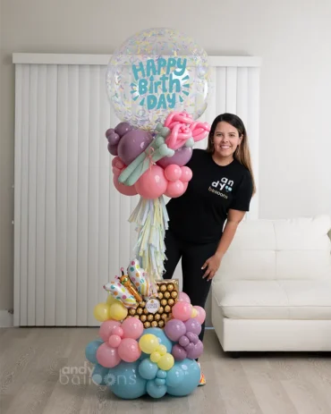 Tall luxury birthday balloon tower with a clear confetti topper, pink and purple floral balloon accents, and a large box of Ferrero Rocher chocolates at the base.